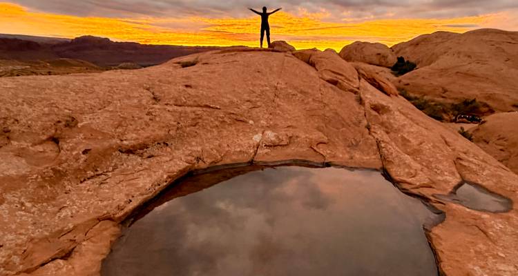 Un voyageur solitaire se tient triomphalement au sommet d'une roche lisse à côté d'une flaque de pluie reflétant le ciel du coucher de soleil aux teintes mélancoliques.