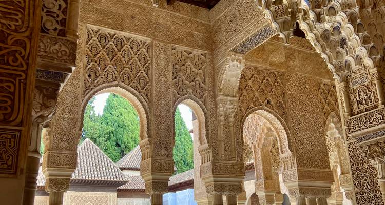 Intricately carved Moorish arches and stucco patterns inside the Alhambra with greenery beyond.