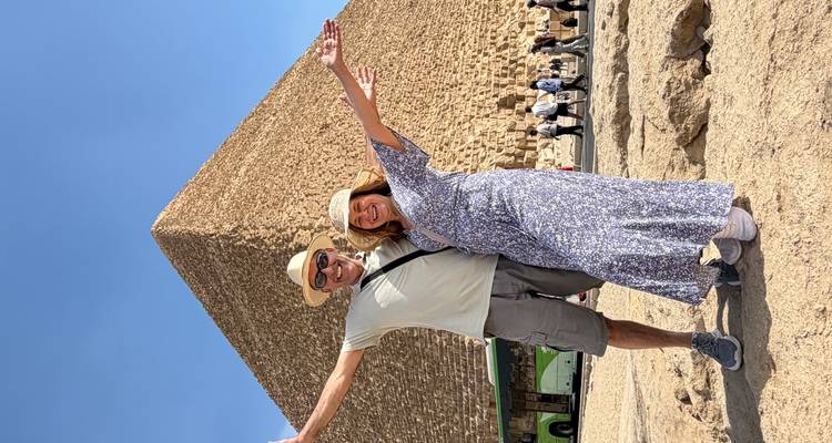 Smiling couple with arms outstretched stand before the Great Pyramid of Giza on a clear blue day.