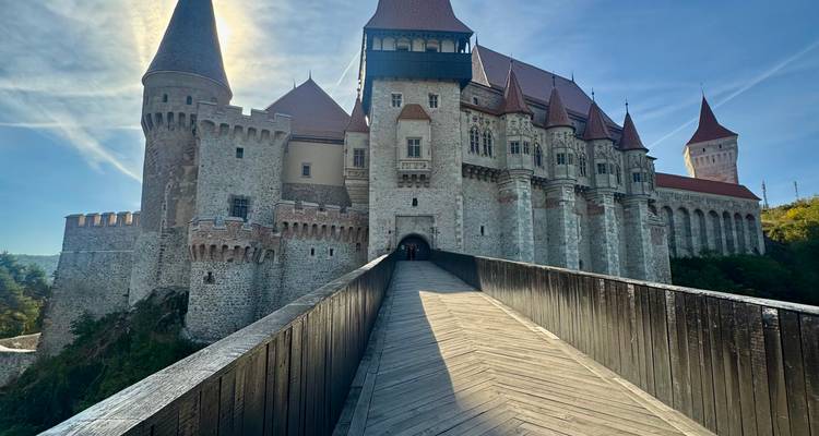 Castillo de estilo gótico de piedra con tejados rojos al que se accede por un largo puente de madera en un día soleado