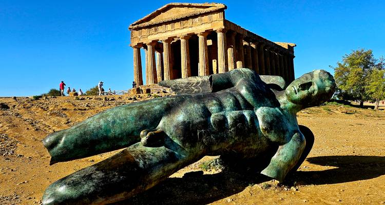 Bronze fallen Icarus statue lying before the well-preserved Temple of Concordia at the Valley of the Temples on a sunny day.