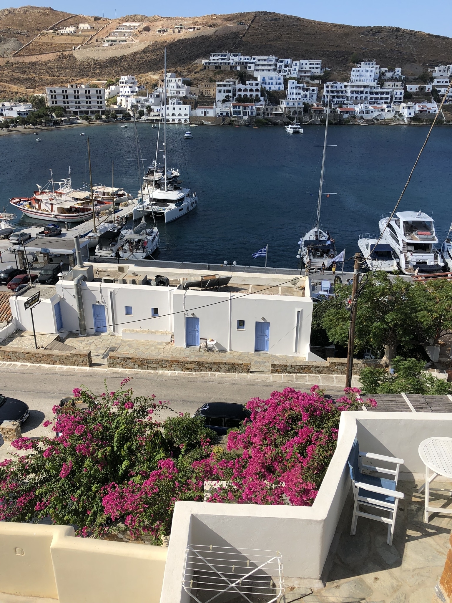 White buildings with blue doors near a dock with boats and a Greek flag.