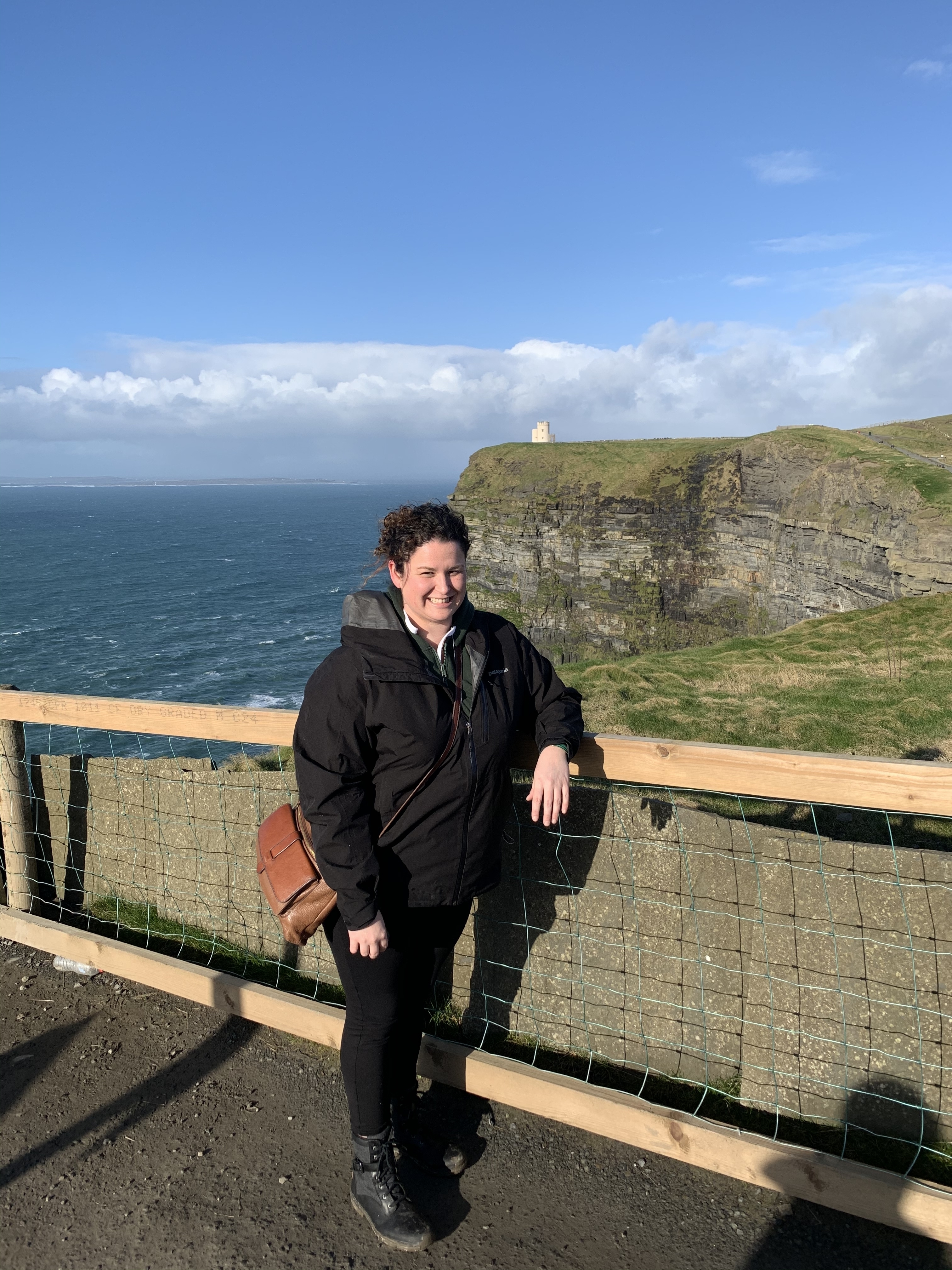 Person standing by Cliffs of Moher under a cloudy sky.