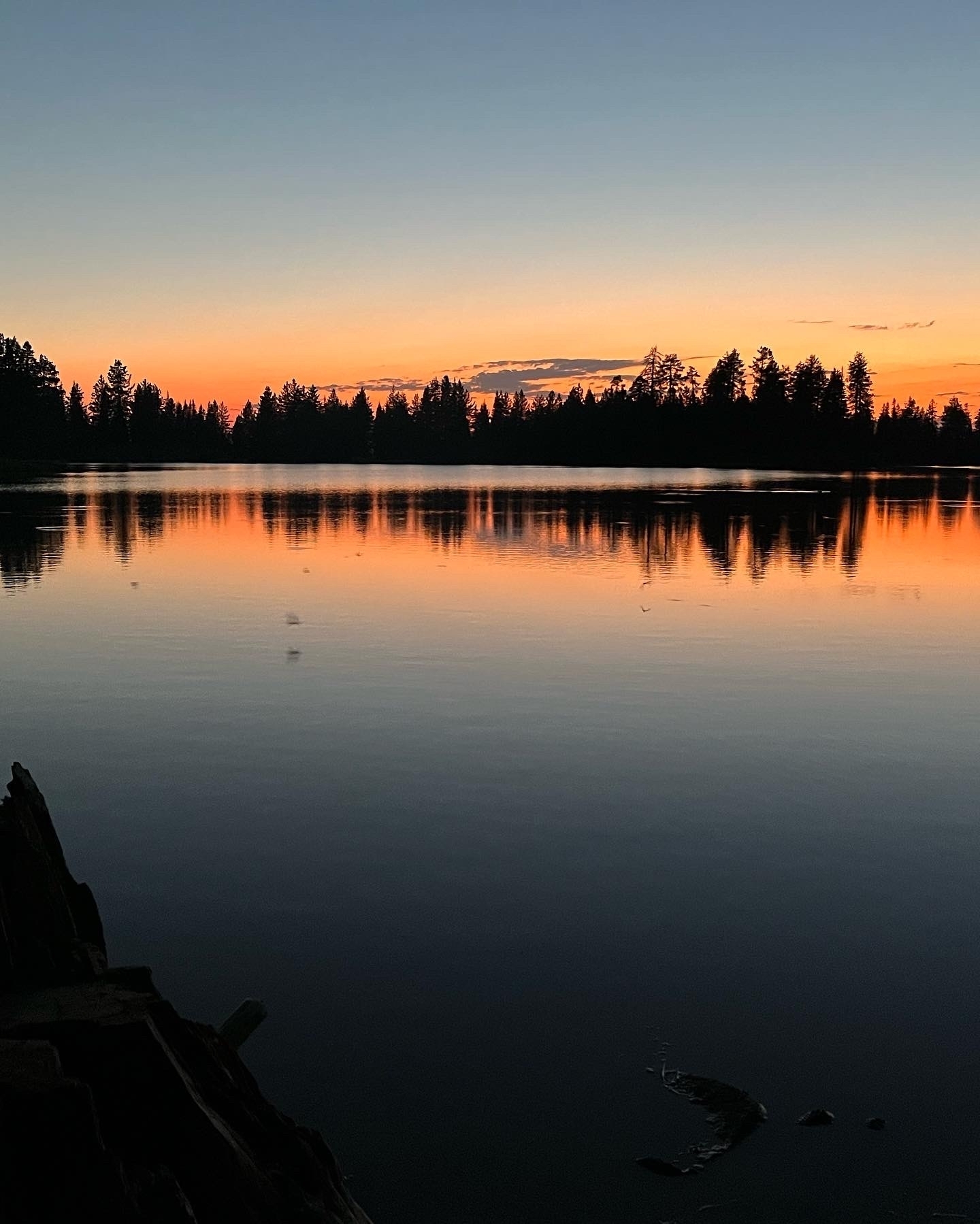 Sunset over a calm lake with reflective water and silhouettes of trees.