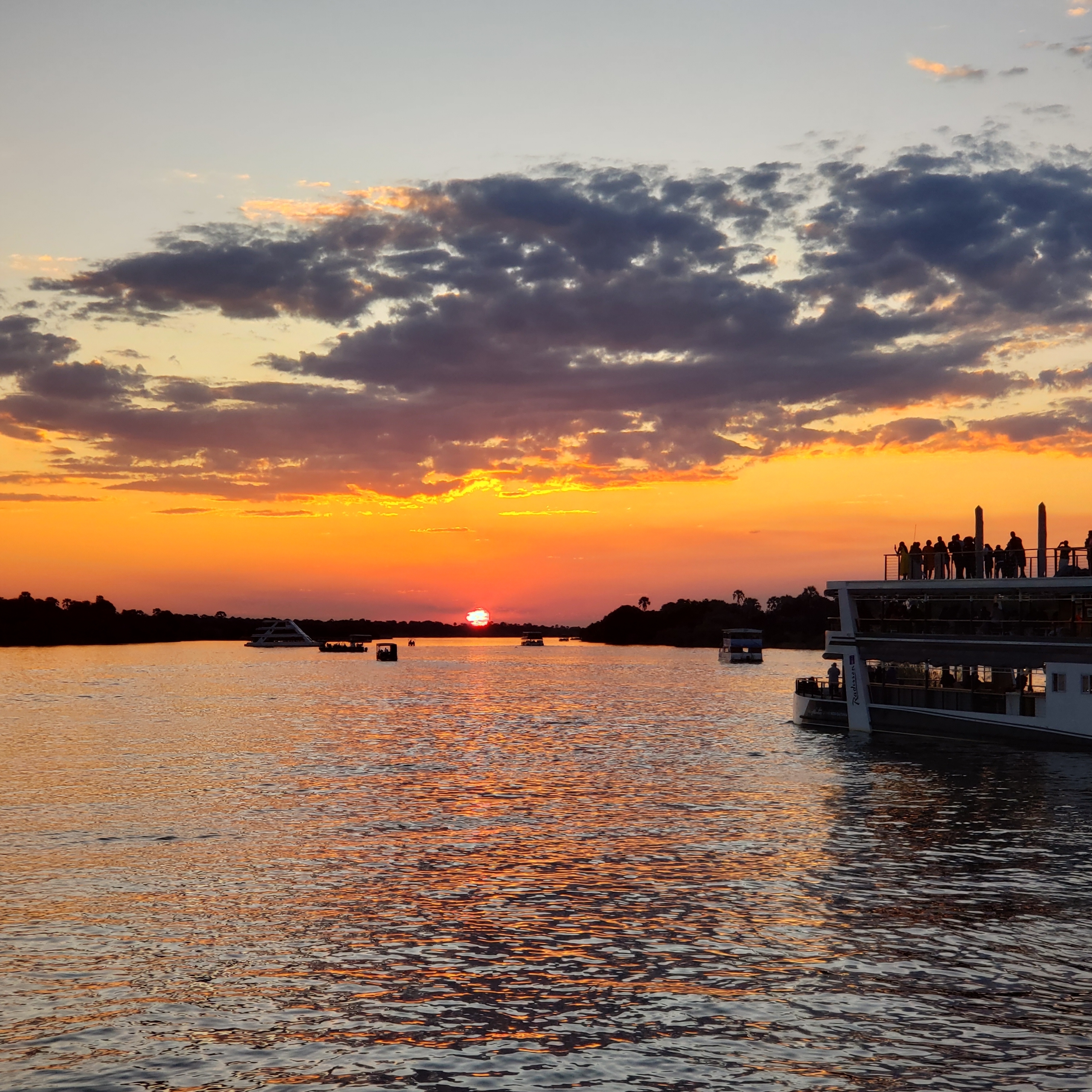 Sunset over a river with boats and silhouettes of people on deck.