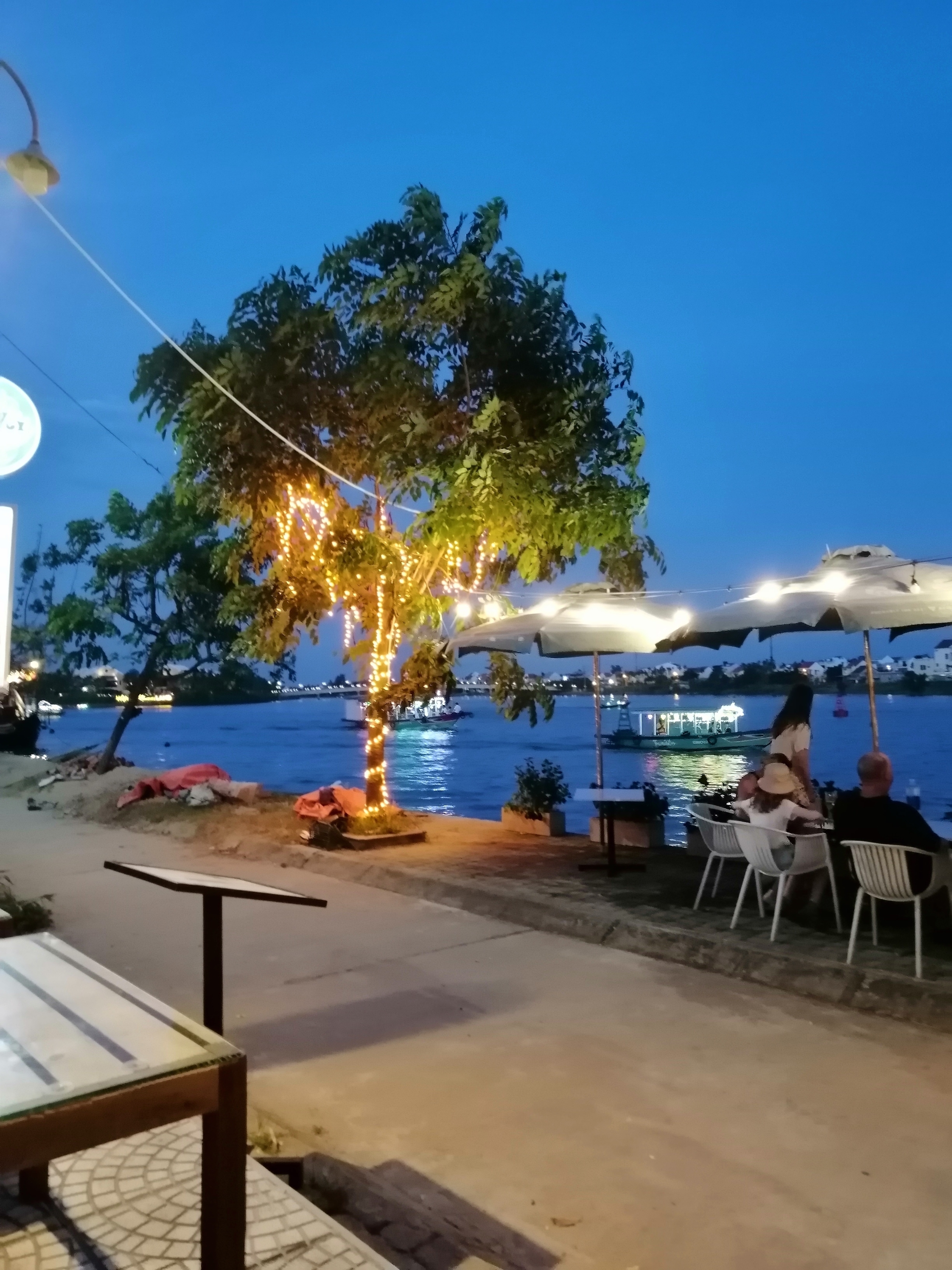 Outdoor dining by a river with lit up trees at night.
