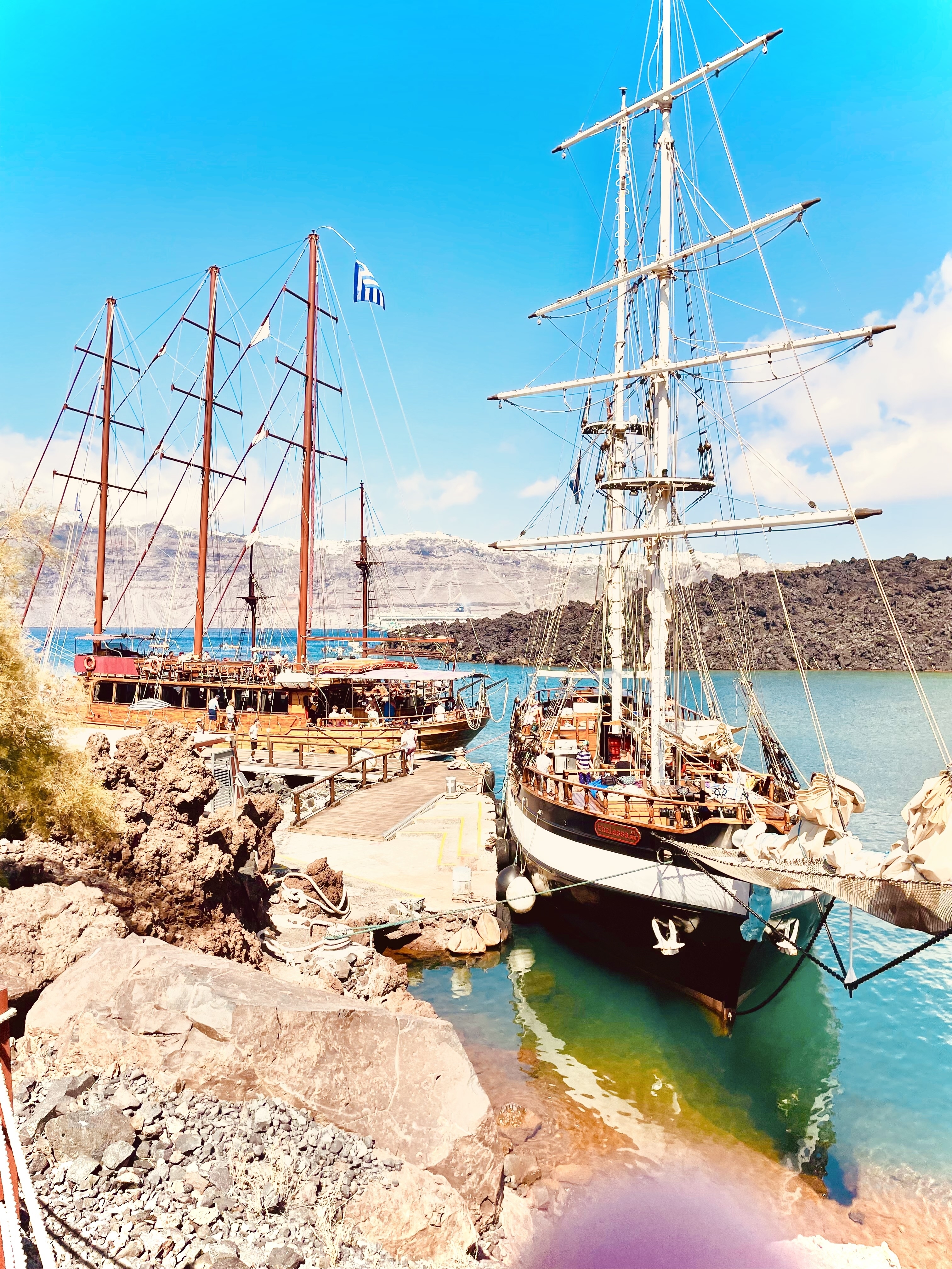 Wooden sailing ships docked at a harbor with a rocky backdrop.