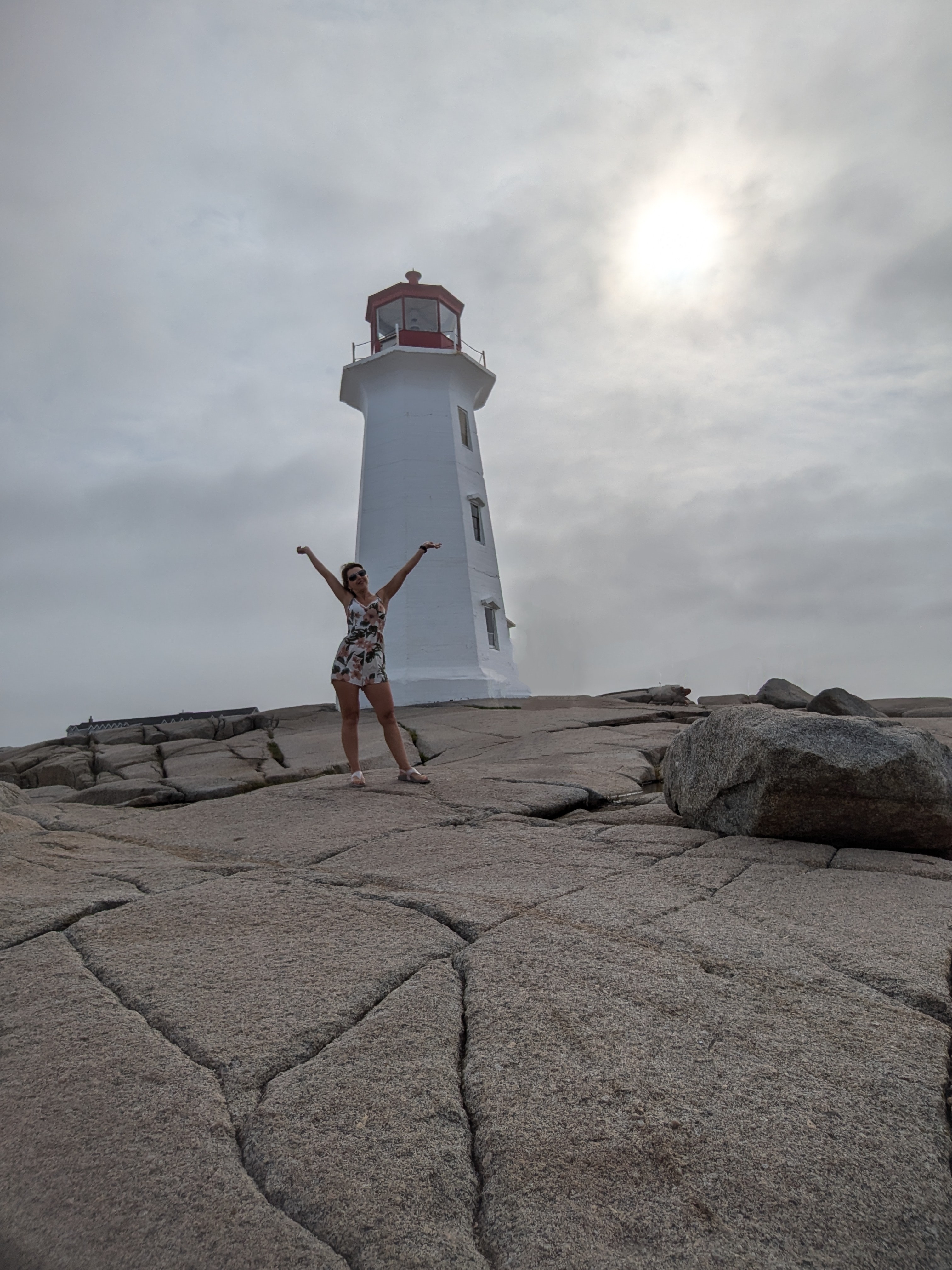 Person posing with raised arms in front of a lighthouse on a rocky area.