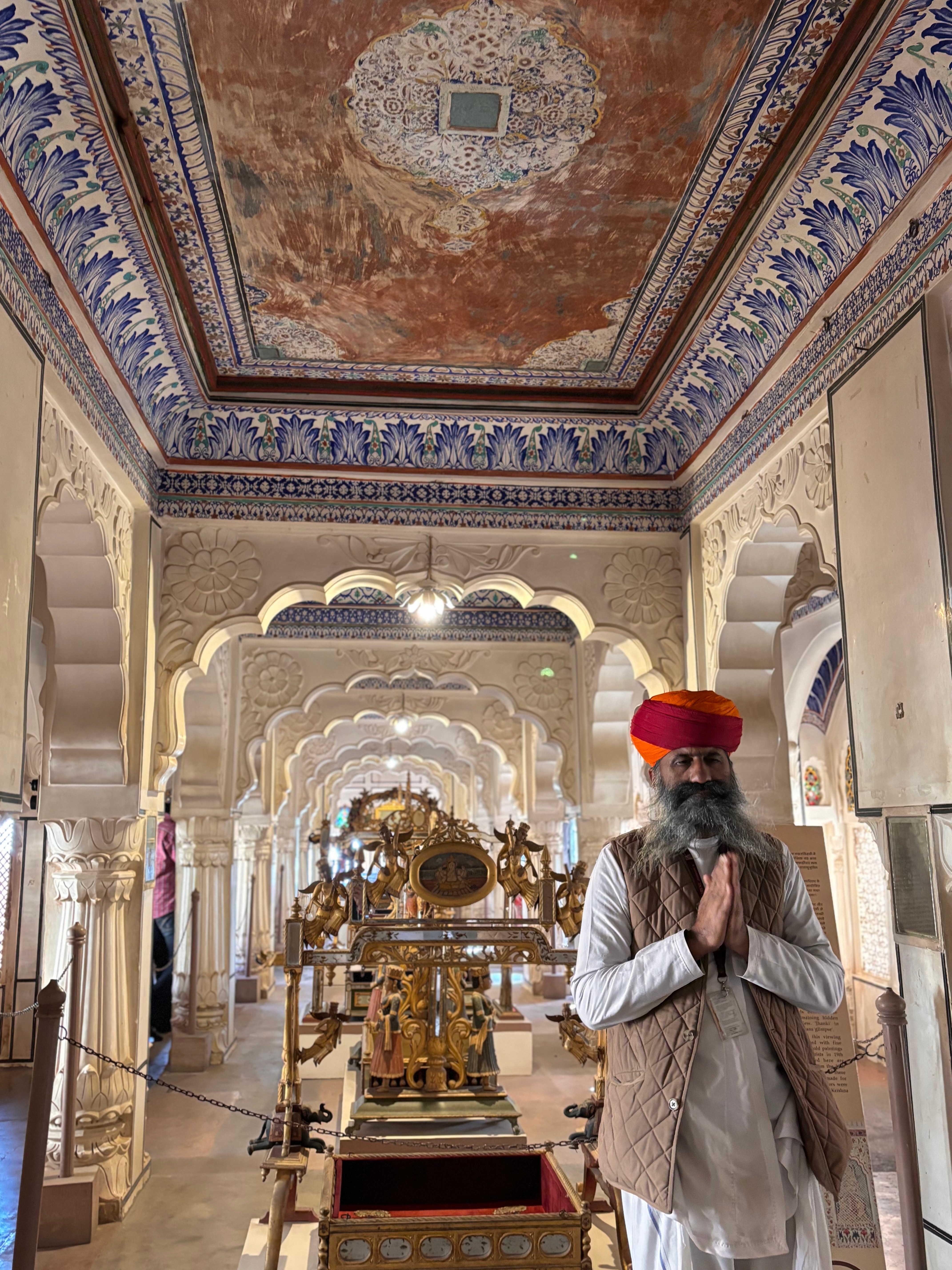 Man in traditional attire standing inside an ornately decorated palace.