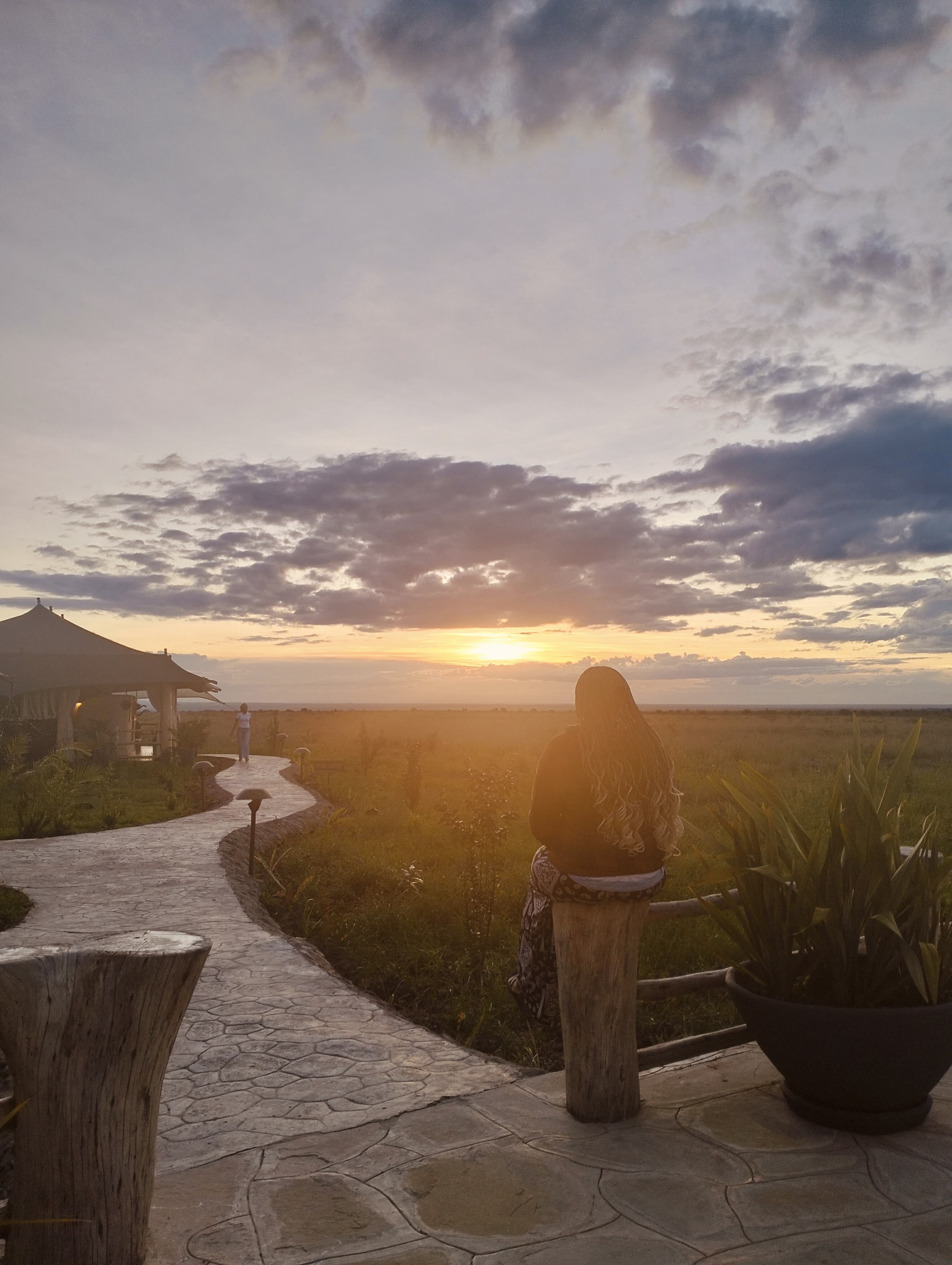 A person looking at a sunset over a grassy plain, with a tent-like structure to the left.
