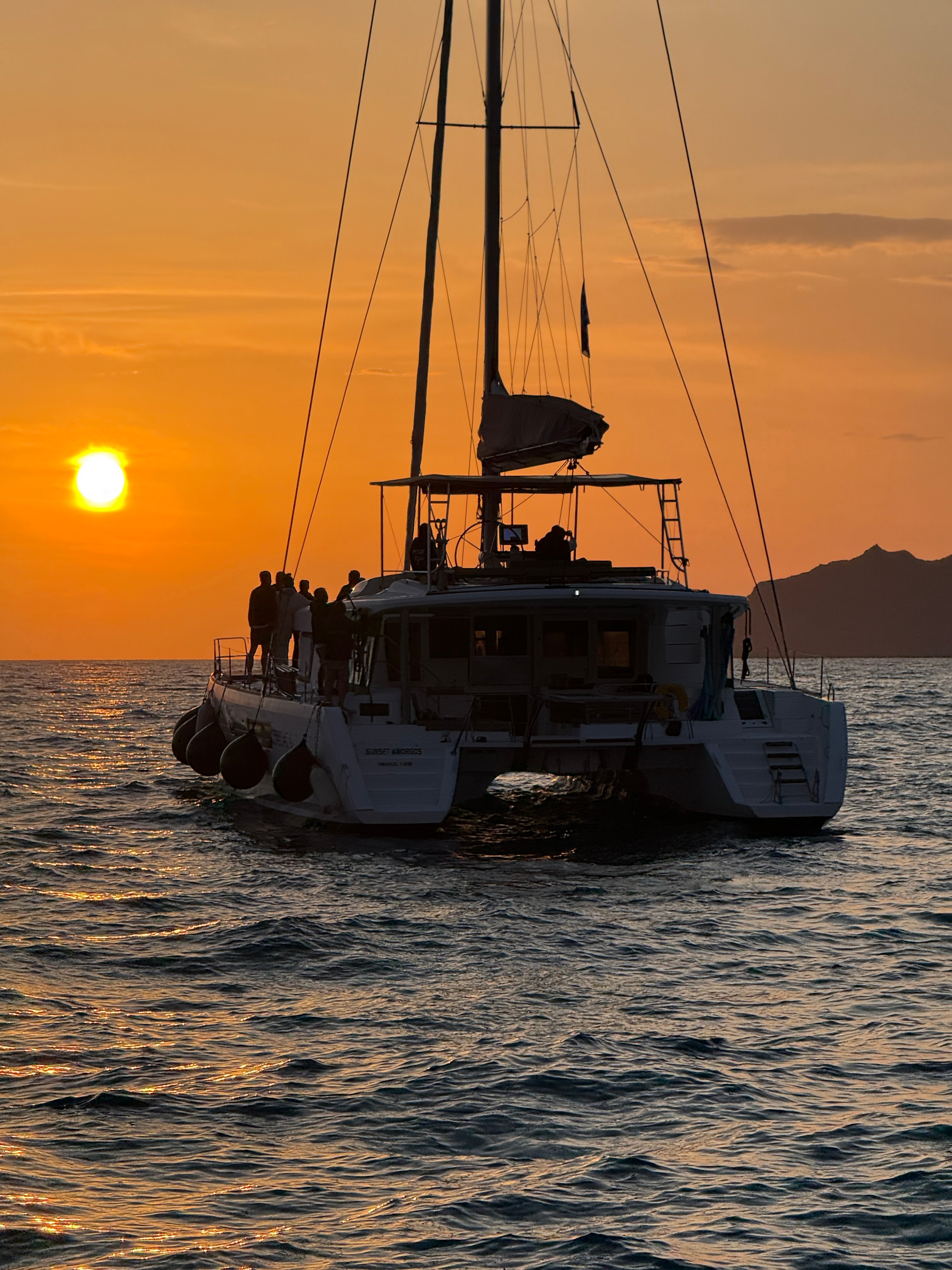 Silhouetted catamaran at sunset with people on board.