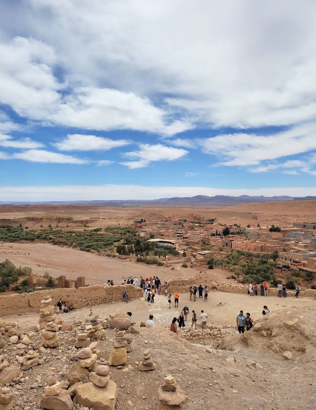 Panoramic view of an arid landscape with a cluster of buildings and people, likely Ait Benhaddou, Morocco.