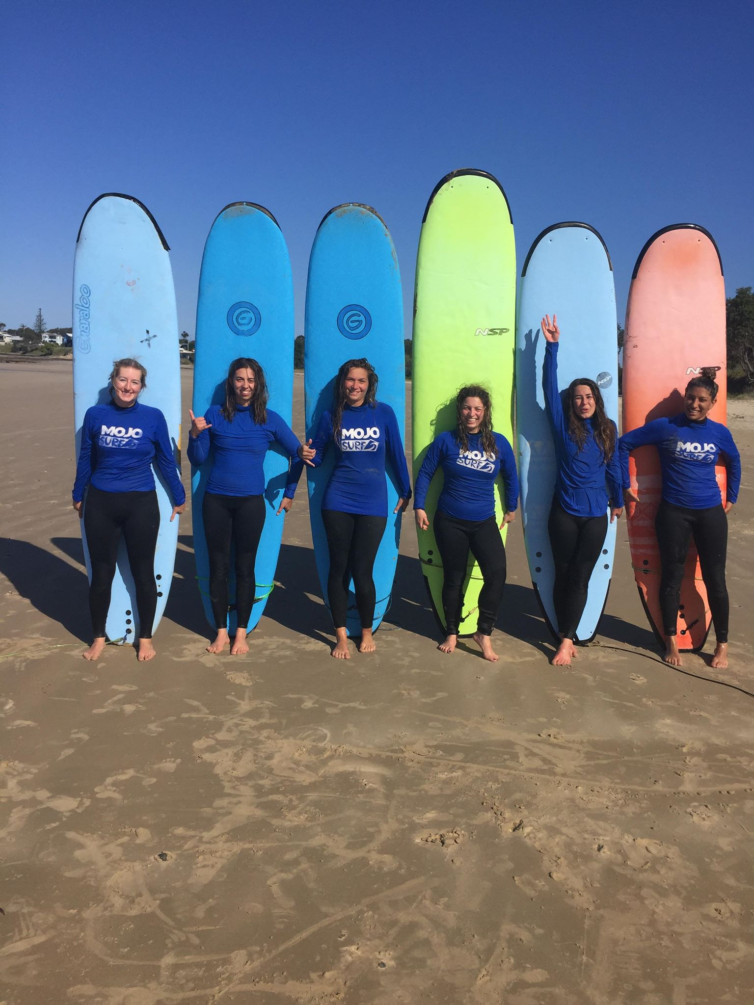 Six surfers in matching uniforms posing with surfboards on a beach.