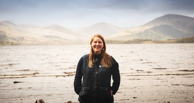 Femme debout près d'un loch avec des collines au loin.