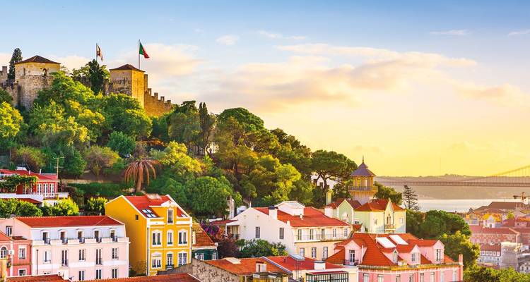 Colorful Lisbon hillside with São Jorge Castle, pastel houses and glimpse of the 25 de Abril Bridge glowing in warm sunset light.