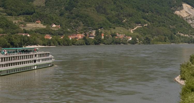 River-cruise ship gliding along a broad green river flanked by forested slopes and scattered village houses.
