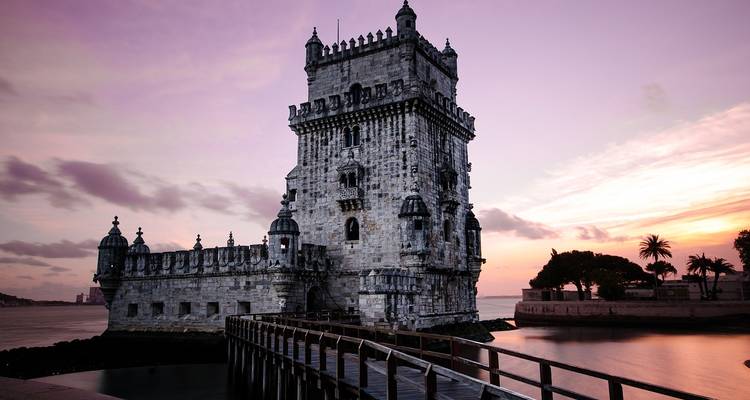 Belém Tower standing at the Tagus River’s edge silhouetted against a pastel sunrise sky with a wooden walkway in the foreground.