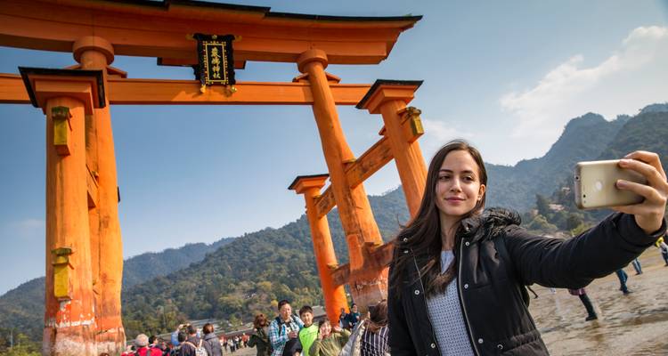 Un viajero sonriente se toma una selfie frente a la brillante puerta torii de color naranja en la isla de Miyajima.