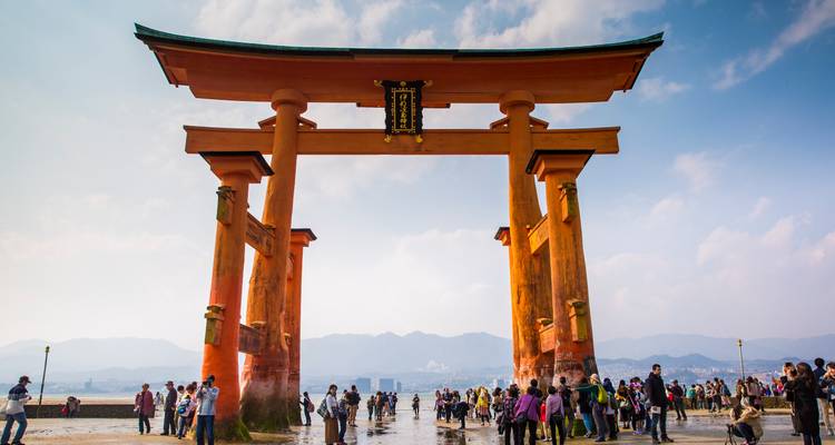 Las multitudes se reúnen en la marea baja bajo la imponente puerta torii naranja con las montañas en la bruma detrás.