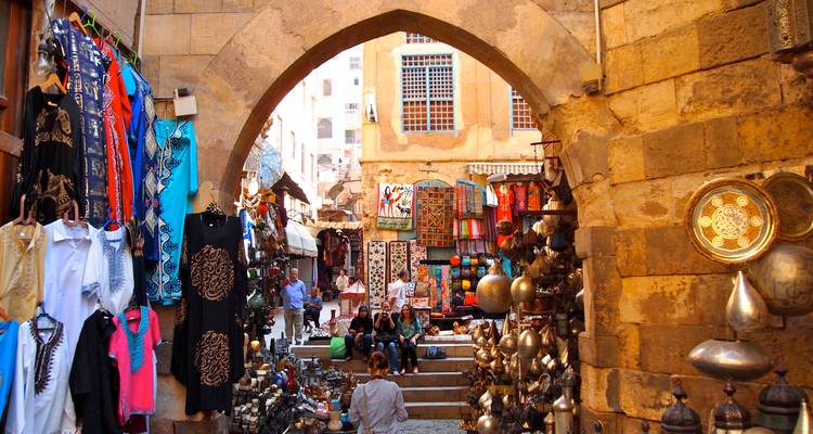 Colorful bazaar alley filled with hanging textiles, brassware and shoppers under a stone arch