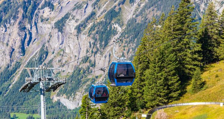 Deux gondoles bleues jumelles voyagent au-dessus de pentes vertes avec des falaises rocheuses abruptes en arrière-plan.