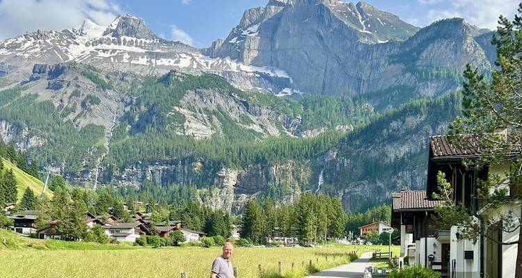 Un homme se tient sur un sentier de village avec de falaises imposantes parsemées de neige s'élevant derrière des chalets.