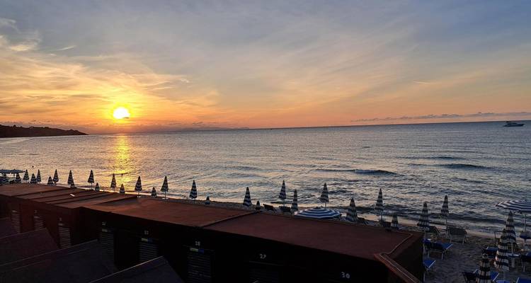 Golden sunset over calm sea with rows of closed beach umbrellas along the shore.