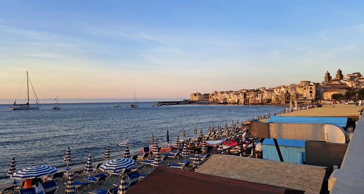 Evening light over a historic coastal town with moored sailboats and beach umbrellas.
