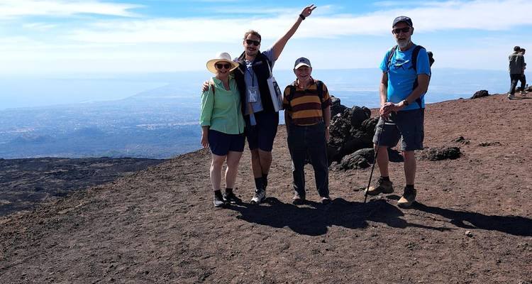 Travelers pose atop the dark lava slopes of Mount Etna with distant coastline below.