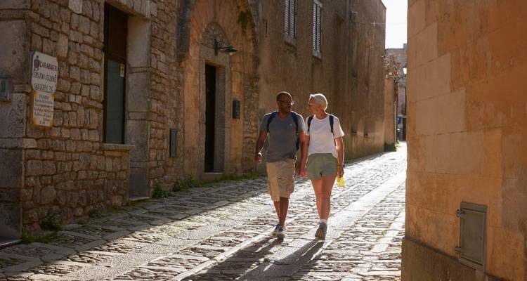 A couple strolls a sunlit cobblestone alley lined with stone buildings.
