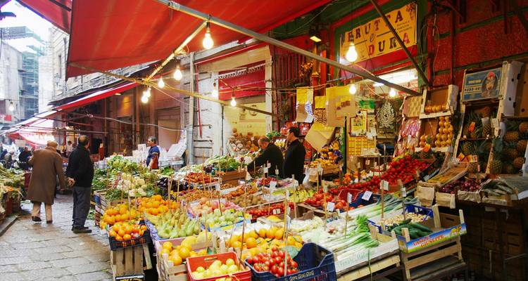 Bustling Sicilian street market bursting with colorful fruit and vegetable stalls under red awnings.