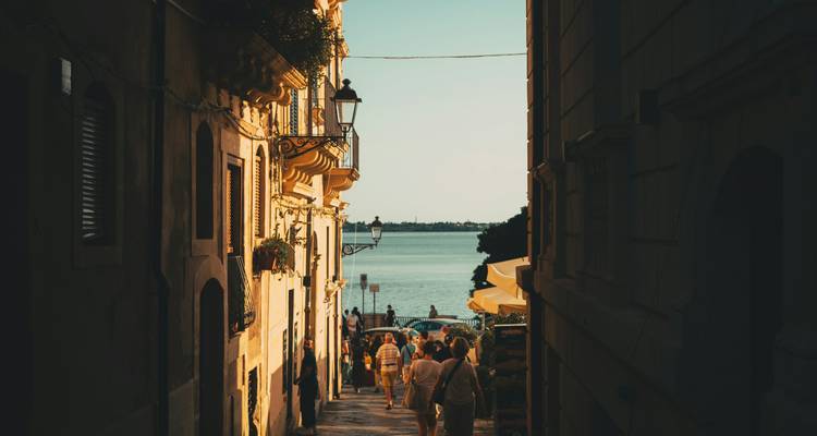 People walk down a narrow sunlit alley toward the waterfront at dusk.