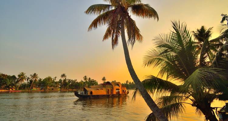 Hausboot segelt bei goldenem Sonnenuntergangslicht auf den Backwaters von Kerala mit silhouettierten Palmen.