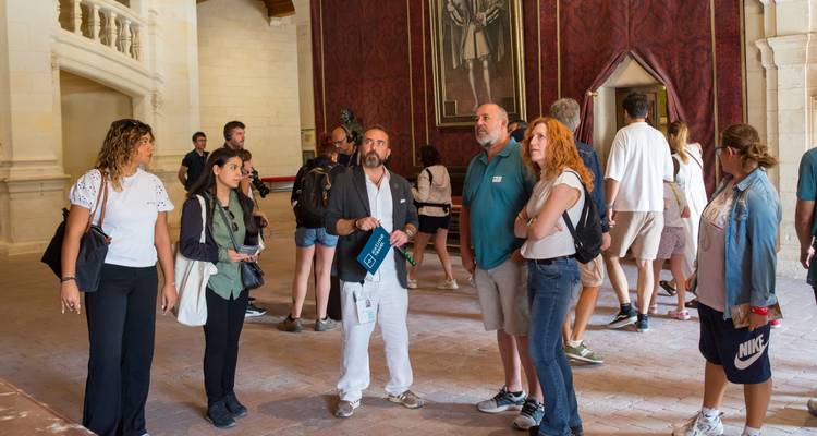Reisegruppe, die in einem dekorierten Saal eines historischen Schlosses mit roten Wänden und Porträts zuhört.