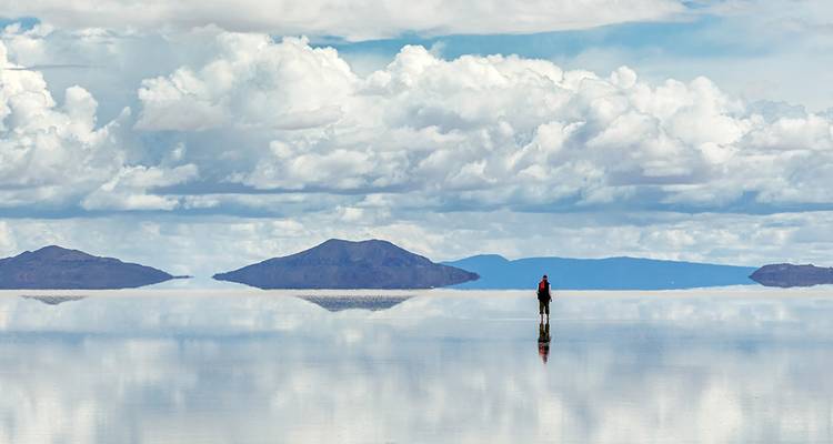 Een solo reiziger die staat op het spiegelachtige Uyuni-zoutvlakte onder dramatische wolken.