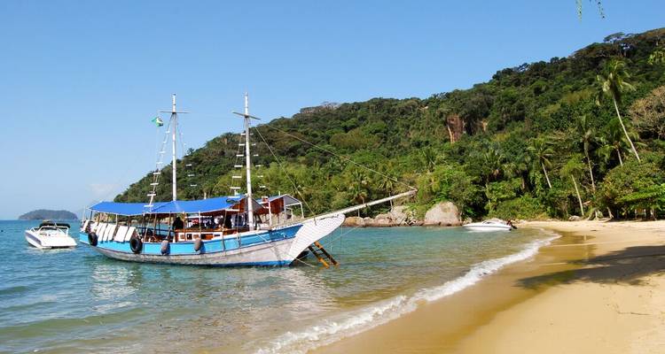 Traditional schooner anchored off palm-fringed sandy beach with clear blue water.