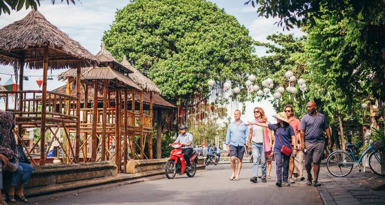 Travelers walk and ride scooters along a lantern-strung street lined with bamboo buildings and lush trees in a historic town.