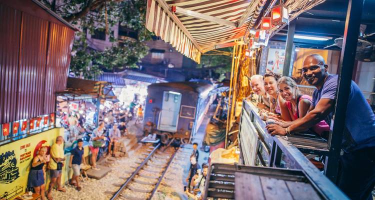 Tourists lean from a café balcony above the famous Hanoi train street as a locomotive passes inches away through a lively night market.