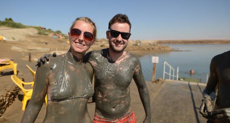 Pareja sonriente cubierta de barro del Mar Muerto posando junto a la orilla.