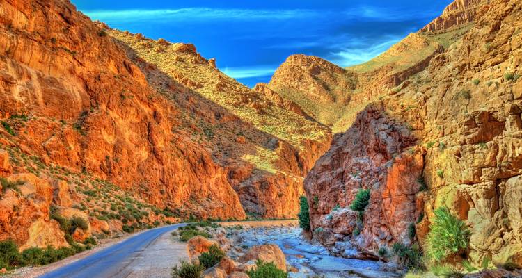 Vivid red canyon walls of Todra Gorge rise above a narrow river and winding road under a deep blue sky
