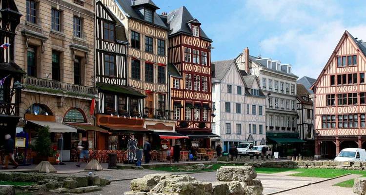Escena medieval de calle con entramado de madera de Rouen con cafés y ruinas de piedra en primer plano.