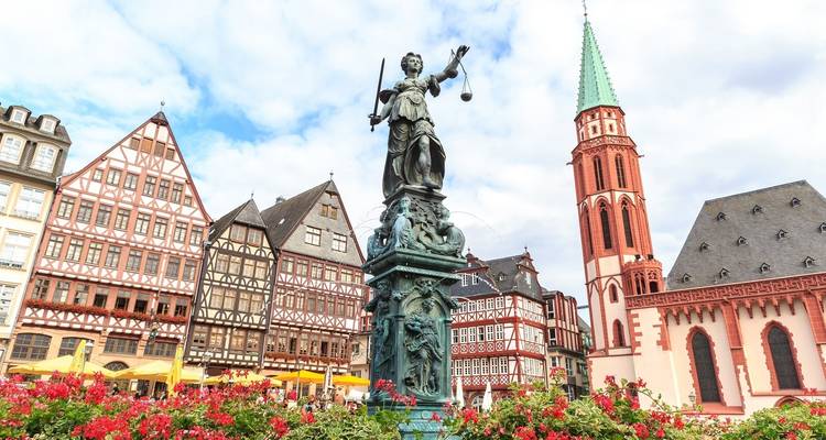 Statue de la Justice sur la Place de la Fontaine avec des bâtiments historiques et un clocher d'église.