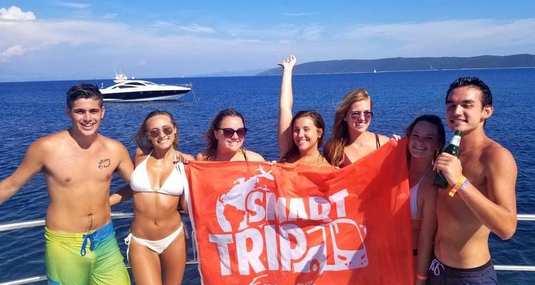 Groupe de jeunes adultes sur un bateau tenant une banderole avec un yacht en arrière-plan.