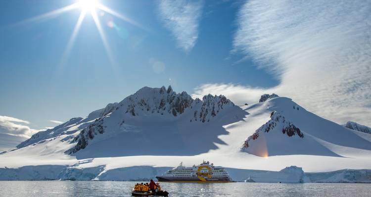 Un pic antarctique ensoleillé s'élève au-dessus d'un navire de croisière tandis qu'un zodiac rempli d'aventuriers traverse des eaux glacées.