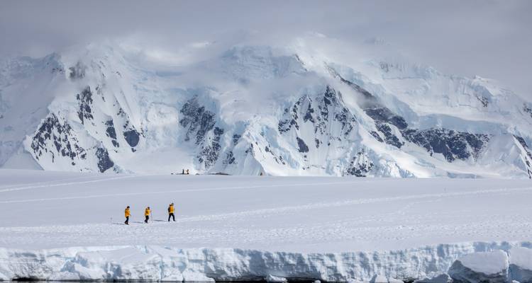 Trois explorateurs vêtus de vestes jaunes traversent un champ de neige sans fin au pied de falaises antarctiques imposantes.