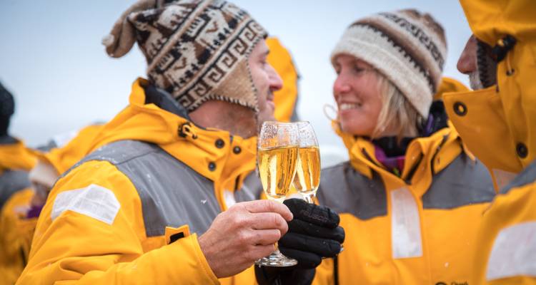 Couple en bonnets tricotés qui font tinter des verres de vin mousseux au milieu de la neige qui tombe sur le pont d'un navire d'expédition.