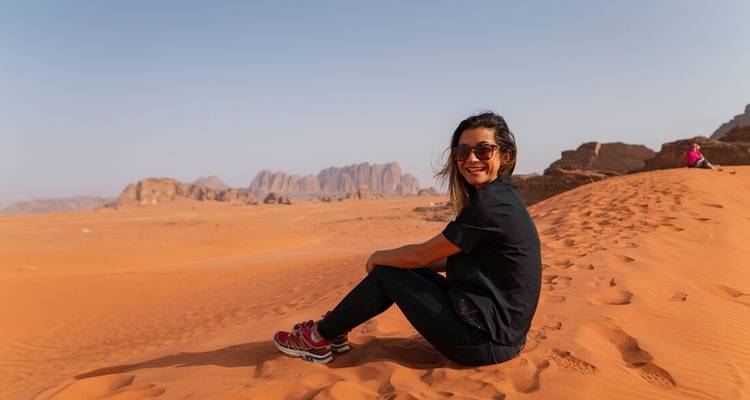 Traveller sits on orange dunes with dramatic sandstone mountains of Wadi Rum in the distance