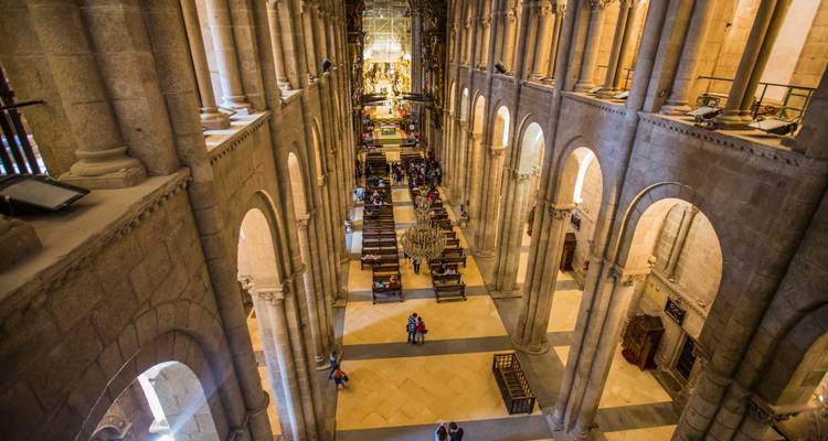 Vista interior elevada del nave y pasillos de una inmensa catedral de piedra con visitantes explorando abajo.