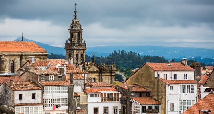 Skyline rústico de Santiago de Compostela con tejados de barro cocido, torre de iglesia y colinas boscosas bajo nubes amenazantes.