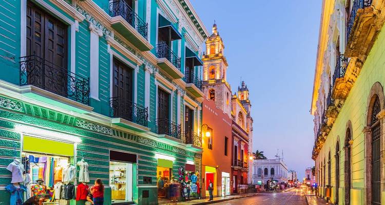 Des bâtiments coloniaux aux façades colorées bordent une rue animée le soir à Mérida avec une cathédrale au loin.